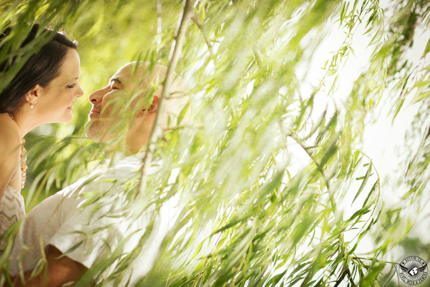 Dark haired lady with pearl earring and beaded  necklace and shirt with chevrons looks into the eyes of a guy with a shaved head in a short sleeve white shirt partially obscured by willow tree branches with lots of soft green leaves all around then at Butler Park near the pond in this breathtaking engagement pic in downtown Austin. 